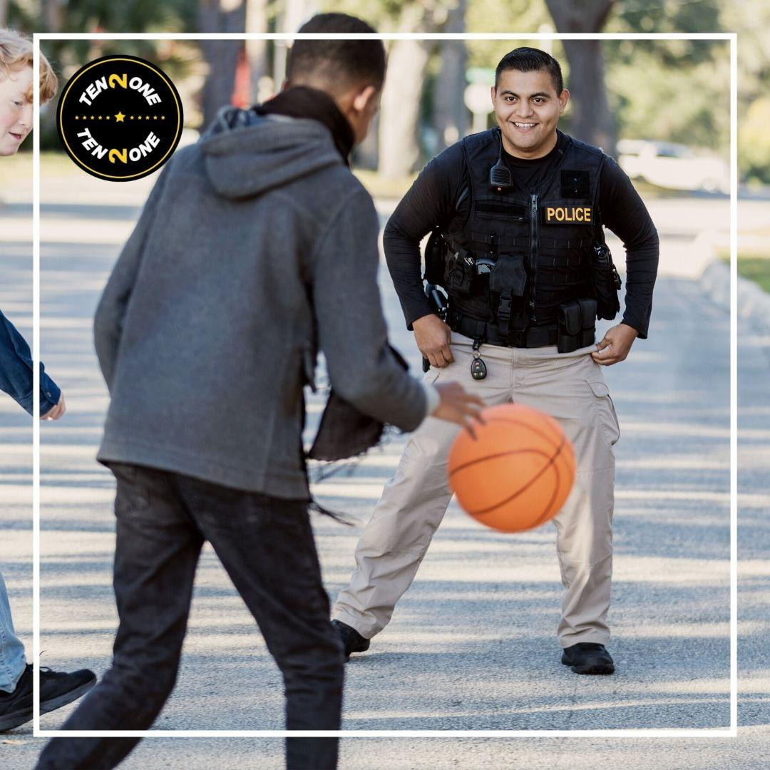 A person in a hoodie dribbles a basketball towards a police officer in tactical gear on a sunny street lined with trees A person in a hoodie dribbles a basketball towards a police officer in tactical gear on a sunny street lined with trees